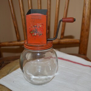 May include: Vintage red and black hand crank food grinder with a glass jar. The grinder has a floral design and the text "Victor" on the side.