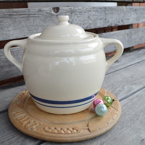 May include: A white ceramic crock with a blue stripe design and a lid. The crock is sitting on a wooden base with a carved design.