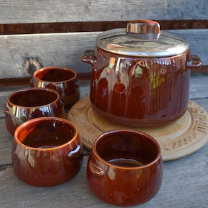 Pode incluir: Um conjunto de cinco tigelas de cerâmica marrom, incluindo uma panela grande com tampa de prata, sobre uma base de madeira. As tigelas têm bordas arredondadas e pequenas alças.