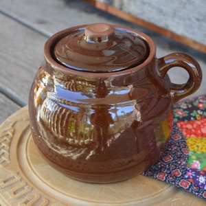 Peut inclure: Un pot en céramique brun avec un couvercle et une anse. Le pot est posé sur une surface en bois avec un tissu patchwork coloré en arrière-plan.