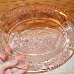 May include: A pink oval serving platter with a floral design. The platter has a raised floral pattern around the rim and a central floral arrangement. The platter is held by a hand.