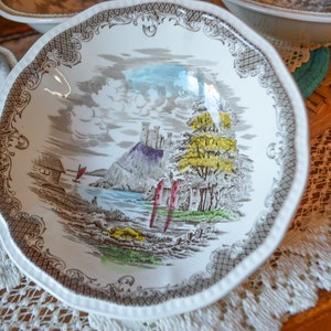 May include: A white ceramic bowl with a brown, gray, and green floral design. The bowl features a castle, trees, and a river. The bowl has a scalloped edge and a decorative border.