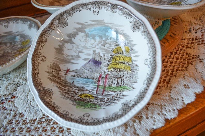 May include: A white ceramic bowl with a brown, gray, and green floral design. The bowl features a castle, trees, and a river. The bowl has a scalloped edge and a decorative border.