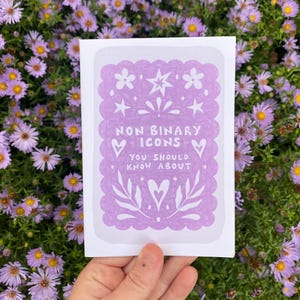Könnte beinhalten: Eine weiße Grußkarte mit lavendelfarbenem Rand und dem Text "NON BINARY ICONS YOU SHOULD KNOW ABOUT". Die Karte wird vor einem Hintergrund aus lila Blumen gehalten.