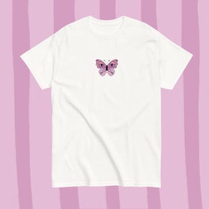 Op de afbeelding: Een wit t-shirt met een klein geborduurd vlinderontwerp in paars, roze en zwart. De vlinder is op de borst geplaatst. De achtergrond is een lichtpaars en wit gestreept patroon.