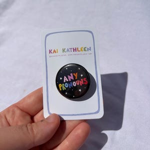 Könnte beinhalten: Ein runder schwarzer Ansteckbutton mit dem Text ANY PRONOUNS in bunten Buchstaben, umgeben von kleinen weißen Sternen. Der Button ist auf einer weißen rechteckigen Präsentationskarte mit dem Text KAI KATHLEEN und einer Website befestigt.