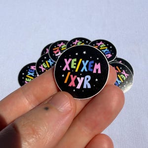 May include: A circular black sticker featuring the text XE/XEM and IXYR in colorful, bold, rounded letters. The background is black with small white stars scattered throughout. The sticker is one of a small pile of identical circular stickers.