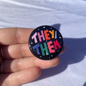 May include: A black button with white stars and the words "THEY/THEM" in rainbow colours.
