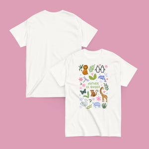 May include: Two white t-shirts on a pink background. One shirt displays a colorful graphic with the text "Nature is Queer" and illustrations of animals including a lion, penguins, and a giraffe. The other shirt is plain.