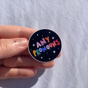 May include: A black and white sticker with white stars. The sticker says "ANY PRONOUNS" in rainbow colors.