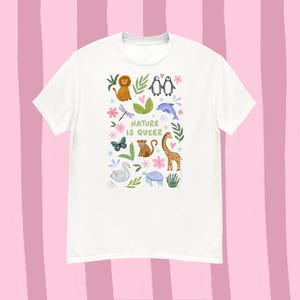 May include: White t-shirt with a colorful illustration of various animals, including a lion, penguins, and a giraffe. The text "NATURE IS QUEER" is printed in a playful font. The design features butterflies, flowers, and leaves.