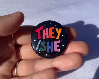 May include: A black button with white stars and the words "THEY/SHE" in rainbow colors.