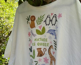May include: A white t-shirt featuring a colorful graphic print of various animals including a lion, penguins, a dolphin, a monkey, a giraffe, a swan, and a turtle, interspersed with leaves, flowers, and hearts, with the text NATURE IS QUEER.