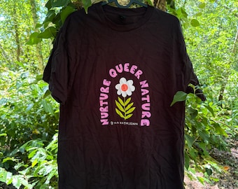 May include: A black short-sleeved t-shirt featuring a graphic print with the text "NURTURE QUEER NATURE" in pink letters arched over a white flower with a red center and green leaves, with the handle "@KAIKATHLEENN" printed below the flower.