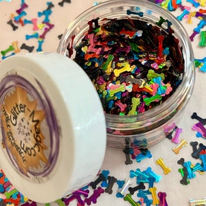 May include: A clear jar filled with colorful iridescent glitter shaped like dog bones. The jar has a white lid with the text "Make Some Magic Glitter" on it.