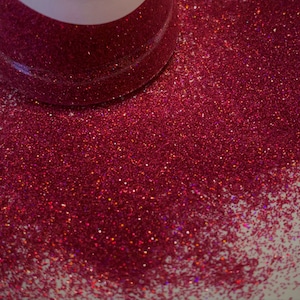 May include: A close-up of a jar of fine red glitter. The glitter is iridescent and has a slight shimmer. The glitter is spilling out of the jar onto a white surface.