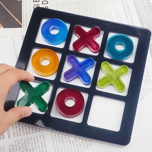 May include: A black tic-tac-toe game board with colorful resin pieces. The pieces are shaped like circles and Xs. The board has nine squares.