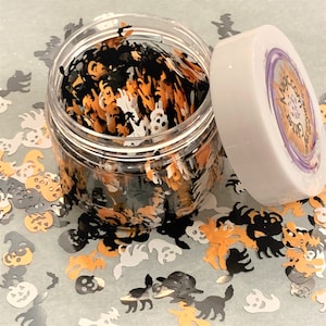 May include: A clear jar filled with black, orange, and white Halloween-themed glitter. The glitter is shaped like ghosts, pumpkins, bats, and skulls. The jar has a white lid with a purple label that says "Magic Sparkle Glitter".