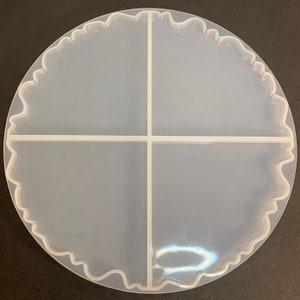May include: A clear, round silicone mold for resin art. The mold is divided into four sections by straight lines and has a wavy, scalloped edge. The mold is designed for creating coasters or decorative pieces.