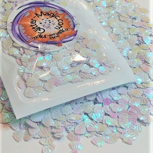 May include: A white plastic bag filled with iridescent white glitter shaped like seashells. The bag has a label that says "Glitter-Magic.com Make your event magical."