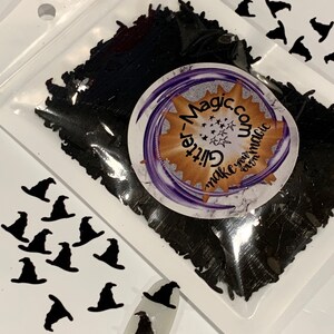 May include: A clear plastic bag filled with black glitter shaped like witch hats. The bag is labeled "Glitter-Magic.com Make your own magic". There are also several black witch hat cutouts on a white background.