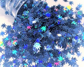 May include: A clear jar tipped over, spilling out a large quantity of blue glitter shaped like cannabis leaves. The glitter has a holographic sheen, reflecting multiple colors. The background is white.
