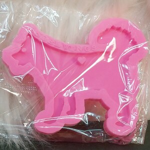 May include: A bright pink silicone mold shaped like a standing dog with a curled tail. The mold features the embossed text DOGWINGS across the upper back area and a small heart shape located near the center of the dog's body, all encased in clear plastic.
