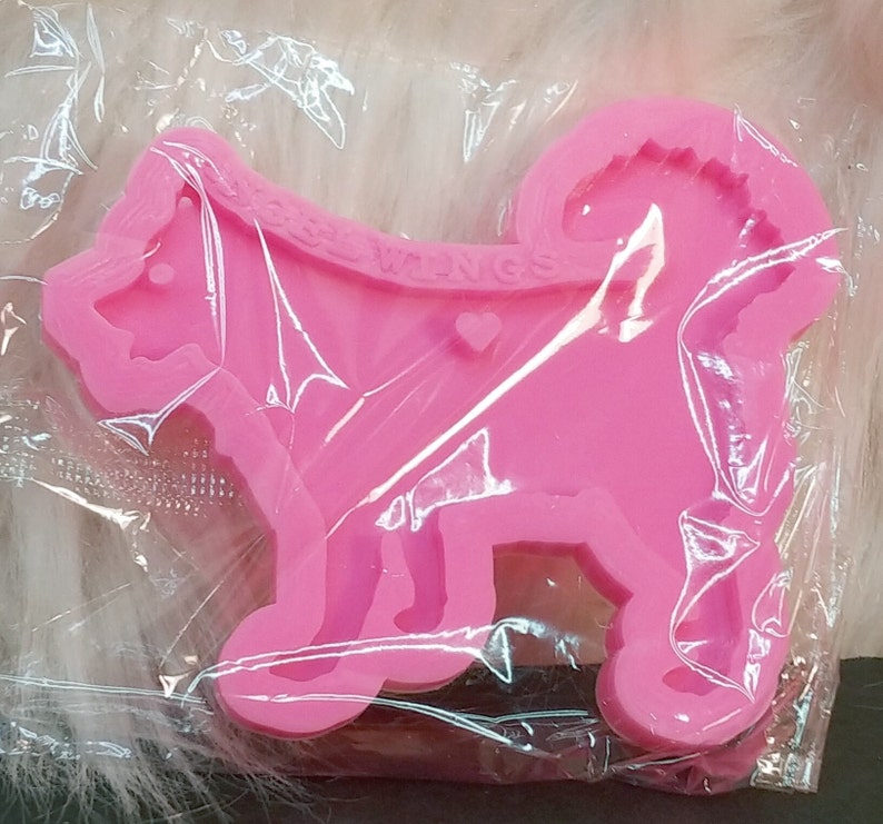 May include: A bright pink silicone mold shaped like a standing dog with a curled tail. The mold features the embossed text DOGWINGS across the upper back area and a small heart shape located near the center of the dog's body, all encased in clear plastic.