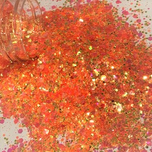 May include: A close-up of orange, gold, and pink glitter. The glitter is loose and scattered on a white surface.