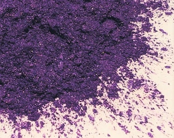 May include: A close-up of a pile of purple glitter. The glitter is scattered on a white surface.