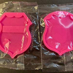 May include: Two pink silicone molds shaped like police badges. One mold has a rectangular space for a name tag and a small heart shape. The other mold is plain.