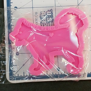 May include: A bright pink silicone mold shaped like a dog with a curled tail, featuring the embossed text "ANGEL WINGS" and a small heart shape on its side. The mold is encased in clear plastic packaging and positioned over a ruler showing inches.