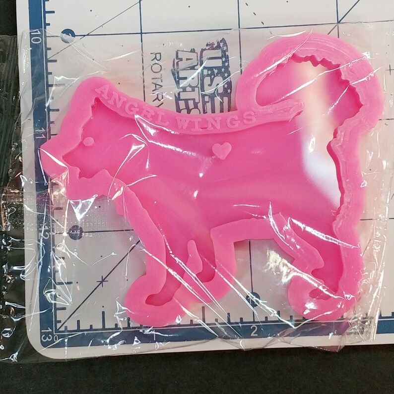 May include: A bright pink silicone mold shaped like a dog with a curled tail, featuring the embossed text "ANGEL WINGS" and a small heart shape on its side. The mold is encased in clear plastic packaging and positioned over a ruler showing inches.