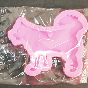 May include: A pink silicone mold shaped like a dog with a curled tail, featuring the embossed text ANGEL WINGS and a small heart shape on the body. The mold is enclosed in a clear, crinkled plastic packaging sleeve for protection and storage.