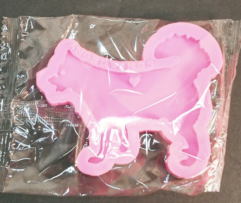 May include: A pink silicone mold shaped like a dog with a curled tail, featuring the embossed text ANGEL WINGS and a small heart shape on the body. The mold is enclosed in a clear, crinkled plastic packaging sleeve for protection and storage.