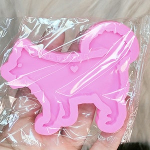 May include: A bright pink silicone mold shaped like a dog with a curled tail, featuring the embossed text "ANGEL WINGS" and a small heart shape on the body. The mold is enclosed in a clear, crinkled plastic packaging sleeve for protection.