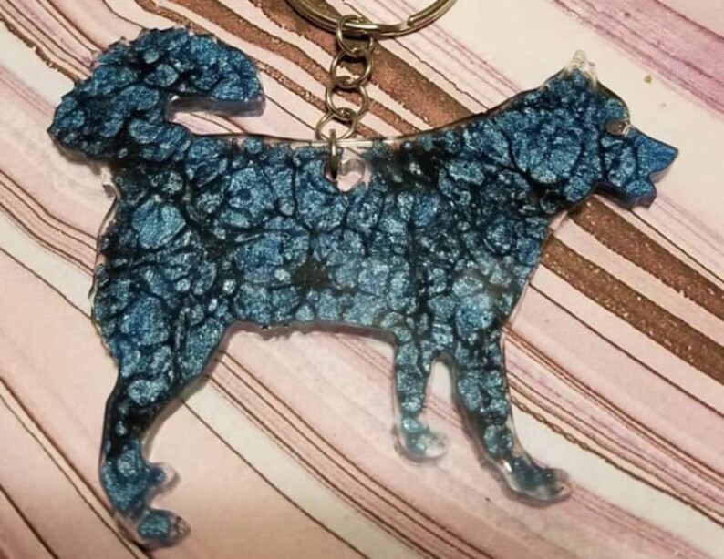 May include: A dog-shaped keychain made of resin with a metallic blue and black marbled finish. The canine silhouette features a textured, swirling pattern of deep blue and dark ink-like veins throughout the body, attached to a silver metal ring.