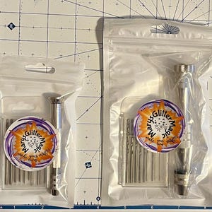 May include: Two clear plastic bags containing silver-colored hand drill tools and drill bits. Each bag has a circular sticker with the text "Marys Glitter Magic make your own" in a purple and orange design. The tools are on a grid background.
