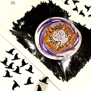 May include: A clear plastic pouch filled with small black witch hat-shaped sequins. A circular sticker on the front features the text Glitter-Magic.com and make your own magic. Several loose black hat sequins are scattered around the pouch.