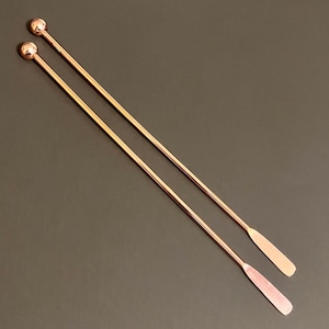 May include: Two gold-colored metal stirring sticks with rounded ends and flat paddle-shaped tips.