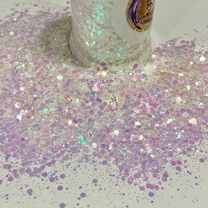 May include: A close-up of iridescent white and purple glitter, with a variety of sizes and shapes. The glitter is scattered on a white surface.
