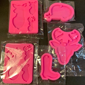 May include: Four pink silicone molds for crafting. The molds are in individual clear plastic bags. The molds are shaped like a deer head, a boot, a camper, and a deer with antlers.