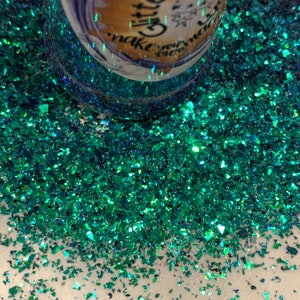 May include: A close-up of teal green iridescent glitter. The glitter is scattered on a white surface. The glitter is chunky and has a holographic finish.