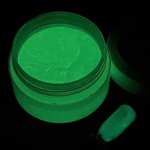 May include: A jar of green glow-in-the-dark nail powder. The powder is in a clear jar with a black lid. The powder is glowing green in the dark.