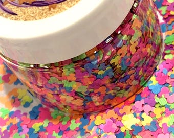 Pode incluir: Um frasco de plástico transparente cheio de confetes de glitter multicoloridos em forma de rato nos tons rosa, azul, laranja e verde. A tampa branca apresenta um rótulo circular com o texto Glitter World e make your own magic.