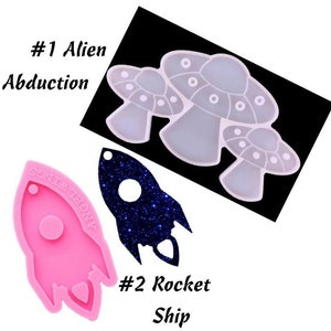 May include: Two silicone molds for resin crafts. One mold is shaped like a rocket ship and the other is shaped like three alien mushrooms. The rocket ship mold is pink and the mushroom mold is white.