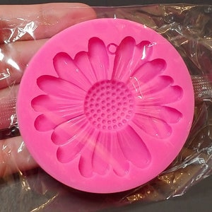 May include: A bright pink silicone mold in the shape of a daisy flower. The mold has a round shape with detailed petals and a textured center. It is designed for crafting and creating decorative elements.