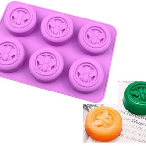 May include: Purple silicone mold with six cavities shaped like bees. The mold is on a white surface. There are three finished soap bars in the image, one green, one orange, and one purple.