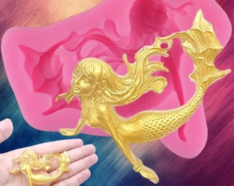 May include: A pink silicone mold featuring a recessed mermaid silhouette and a gold-colored mermaid figurine. The figurine displays a detailed mermaid with flowing hair and a scaled tail, matching the shape of the cavity within the flexible mold.