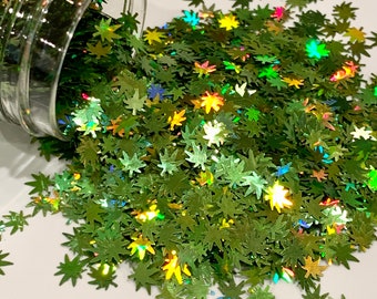 May include: Green iridescent glitter in the shape of marijuana leaves. The glitter is spilling out of a clear glass jar onto a white surface.